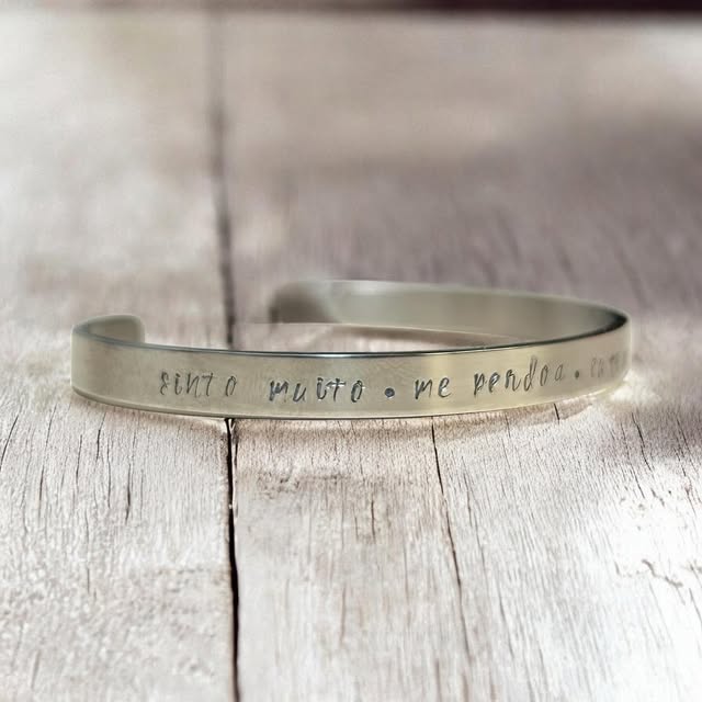 Bracelete de metal gravado à mão com a frase “sinto muito, me perdoa”, sobre madeira clara