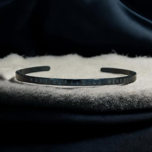 Bracelete gravado com “wabi sabi · dias perfeitos” sobre manta clara e fundo escuro